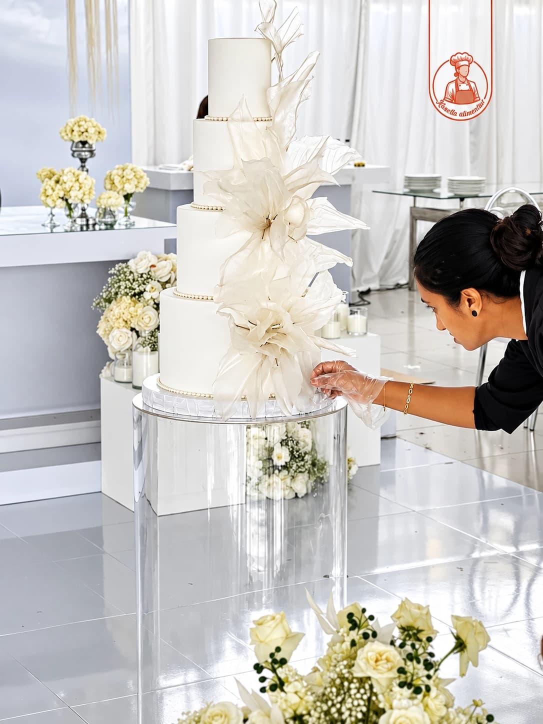 Confeiteira a dar os acabamentos a um bolo de casamento branco com grandes flores de papel de arroz, no local do evento