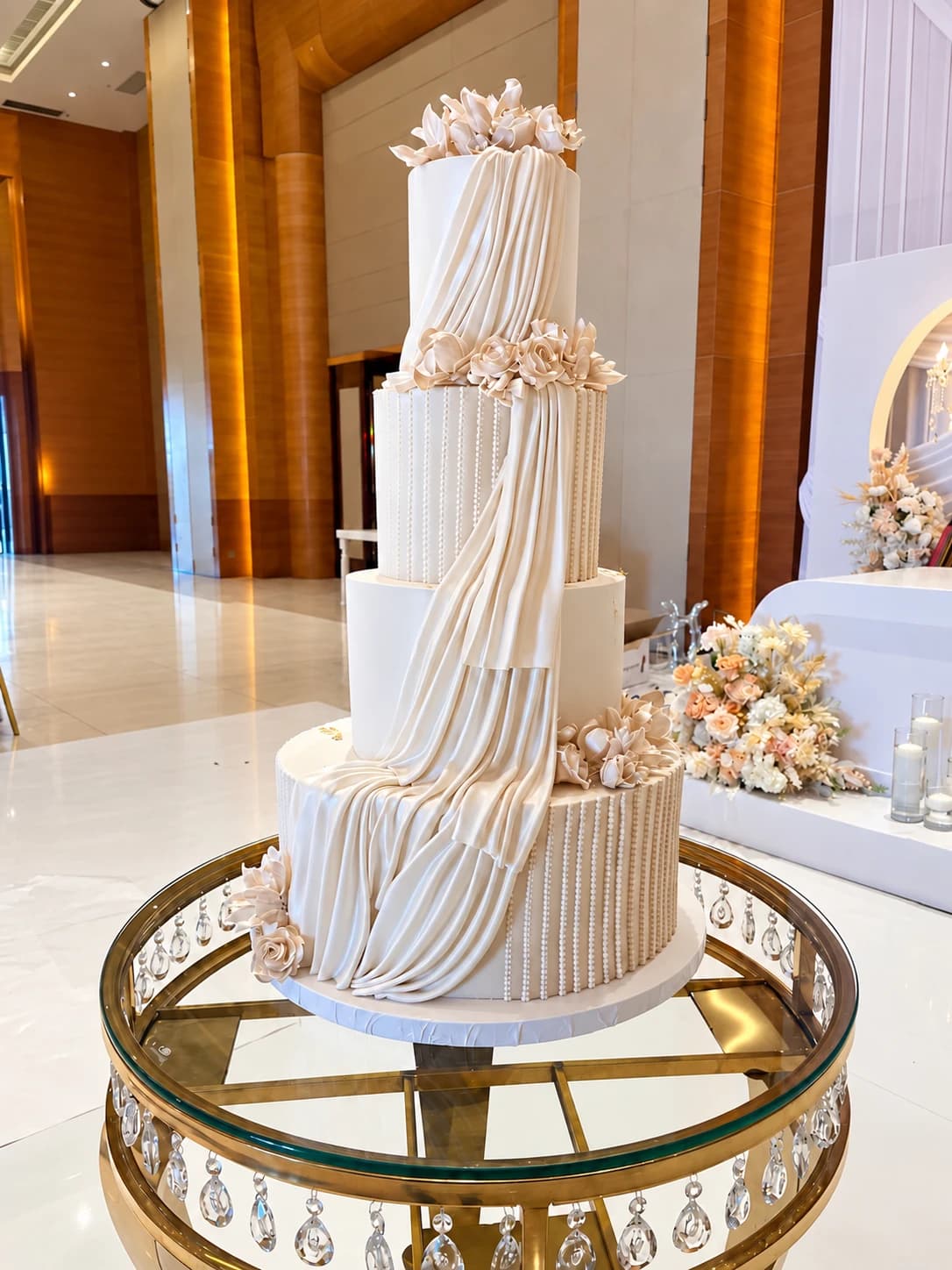 Bolo de casamento de quatro andares em creme, com drapeado de pasta de açúcar e flores delicadas, sobre uma mesa de vidro e dourado