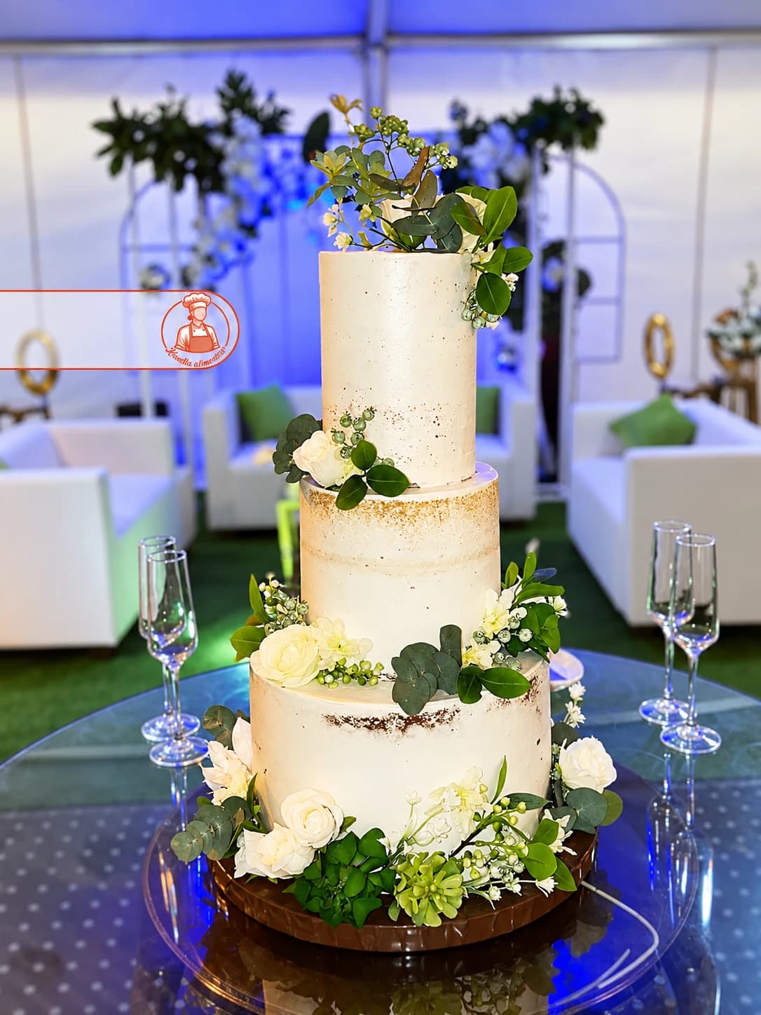 Bolo de casamento de três andares em acabamento rústico, decorado com rosas brancas e folhagem verde, num salão de festa